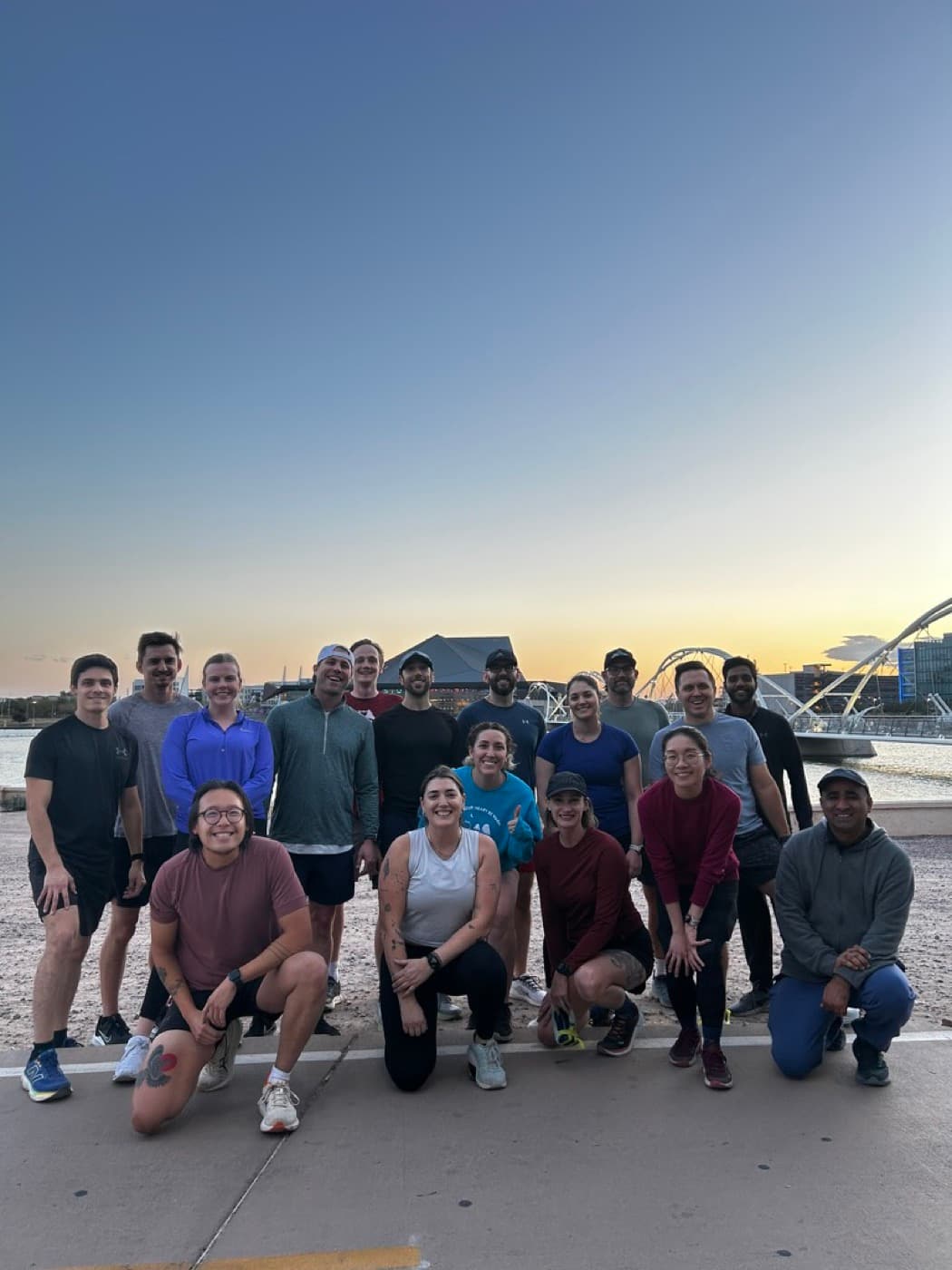 A run club of about eighteen people posing together at Tempe Town Lake at sunrise